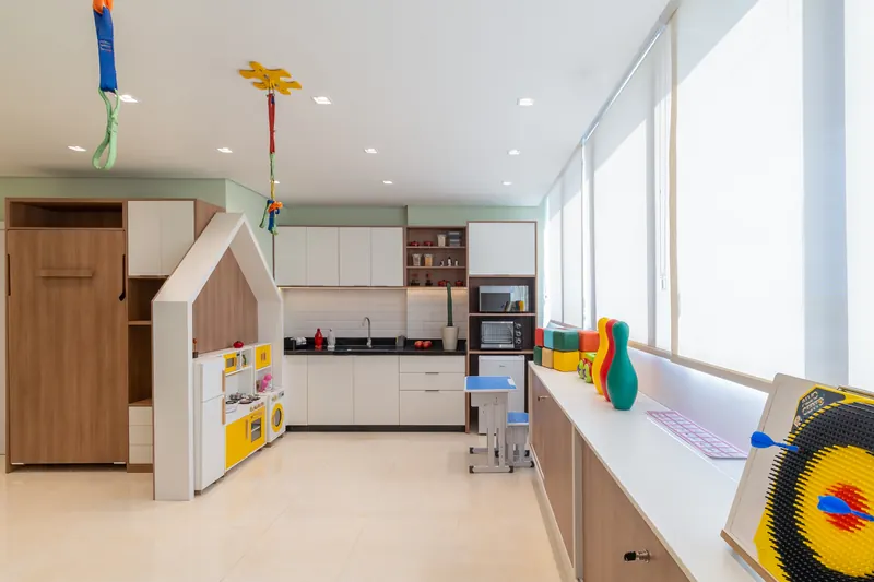 Cozinha com bancada branca e armários de madeira ao lado de uma casinha infantil, em ambiente amplo e iluminado por janelas.