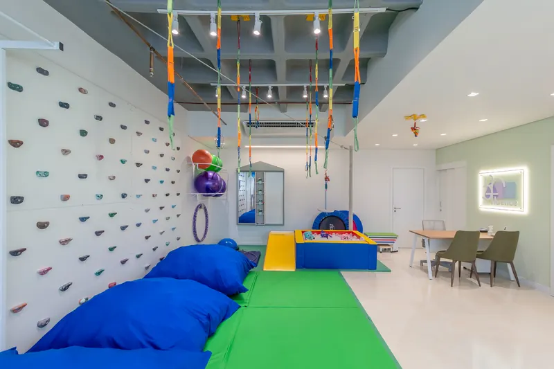 Parede de escalada infantil, balanços e cordas presos à estrutura do teto, sobre tatames coloridos.