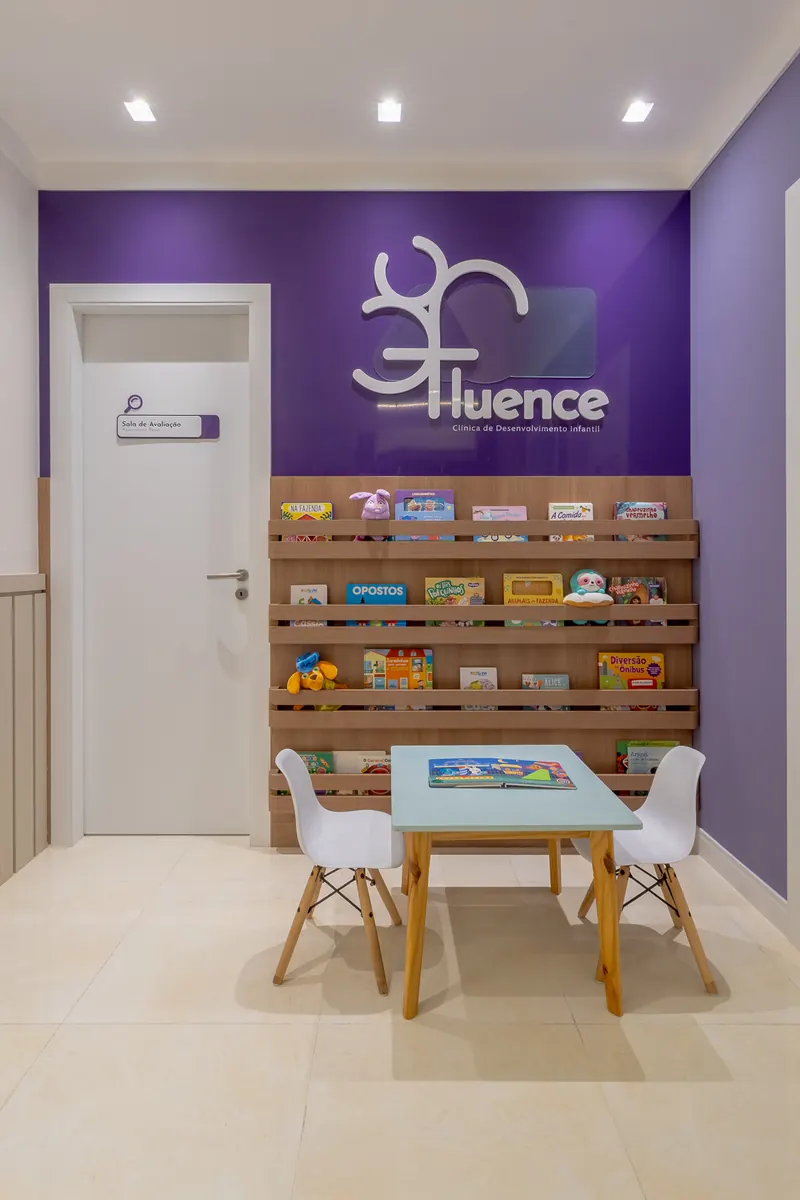 Espera com o logotipo da Fluence na parede roxa, estante de livros infantis em madeira e mesinha com cadeiras brancas.