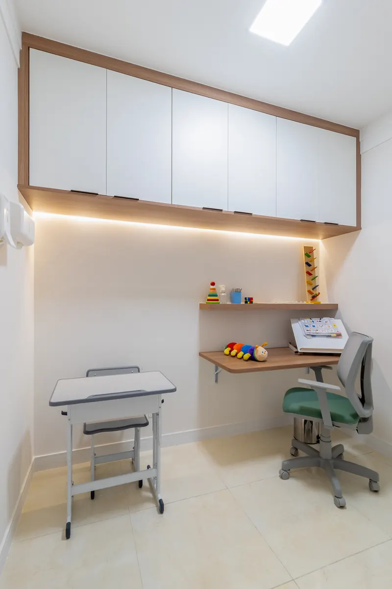 Sala compacta com armários brancos suspensos, bancada de madeira com iluminação embutida, carteira escolar e cadeira ergonômica.