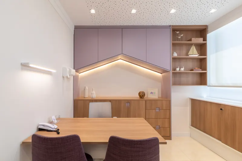 Sala de atendimento com armários lilás em forma de casinha, mesa de madeira e cadeiras ameixa.