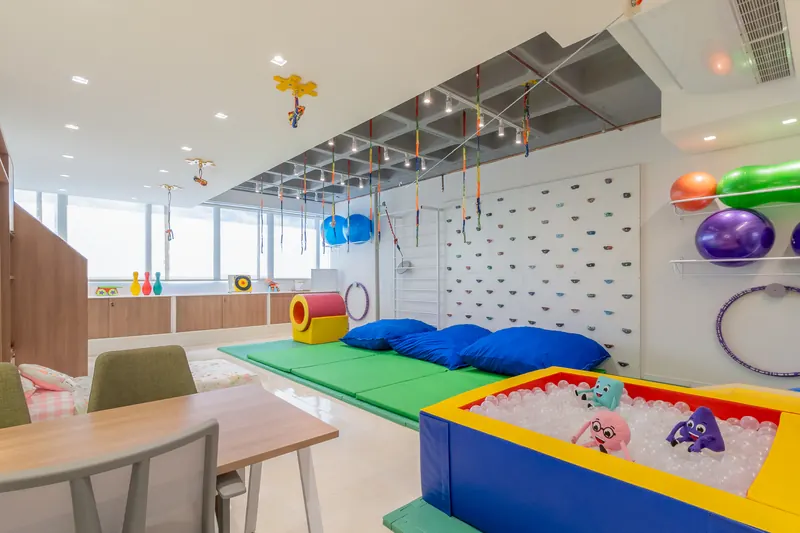 Sala de terapia ocupacional com tatames verdes, piscina de bolinhas, almofadas azuis e equipamentos suspensos no teto.