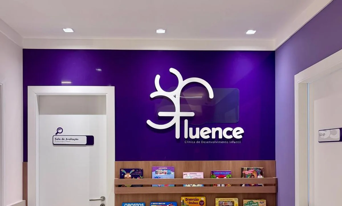 Espera da Clínica Fluence: parede roxa com o letreiro da clínica, prateleiras de livros infantis e a sala de avaliação ao lado.